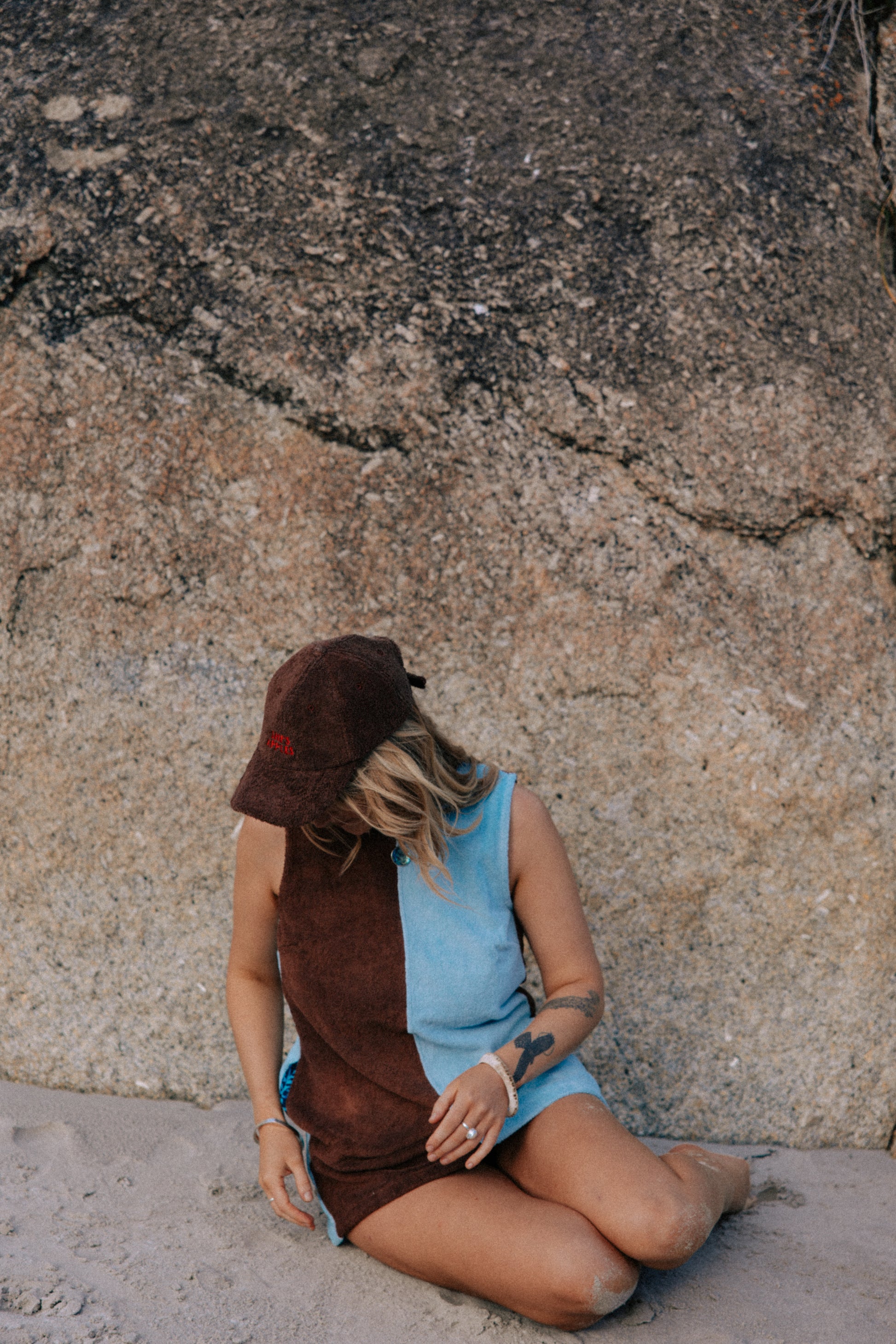 Person wearing a brown hat and blue dress sitting against a rocky wall wearing brown hat.